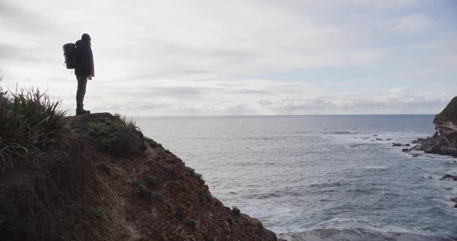 A solo hiker with a backpack reaches the edge of a seaside cliff and gazes out at the ocean