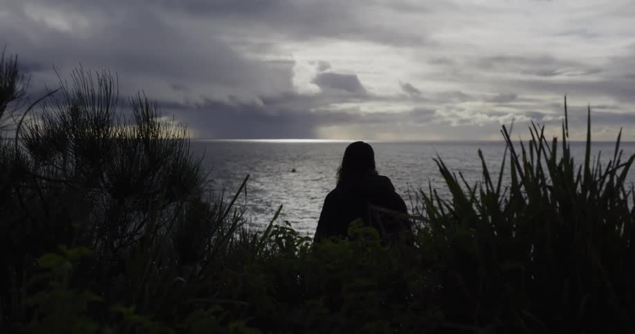 A silhouetted person with long hair sits on a cliff's edge, contemplating the vast ocean under a dark, cloudy sky