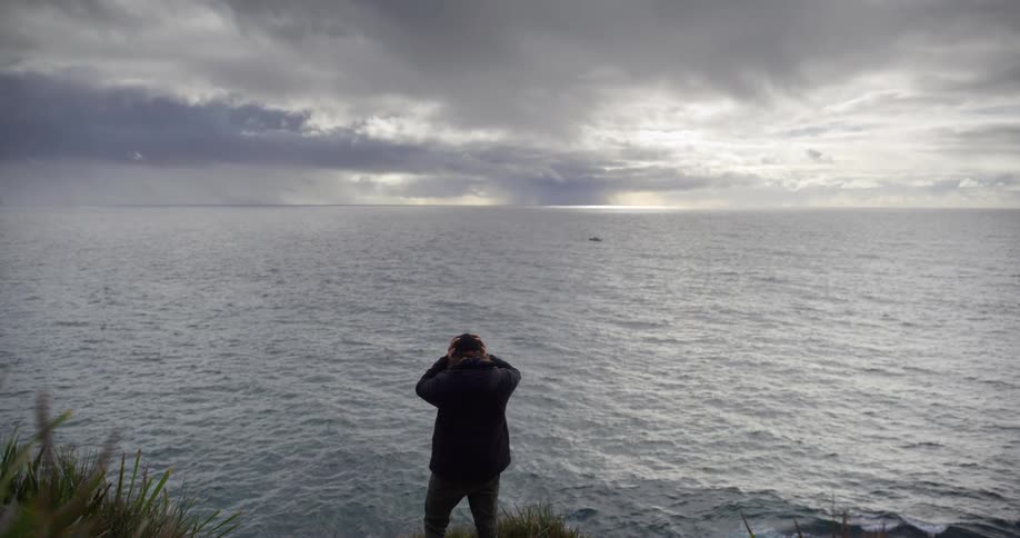 A solitary figure stands on a cliff, overlooking the vast and cloudy ocean as sunlight breaks through on the horizon