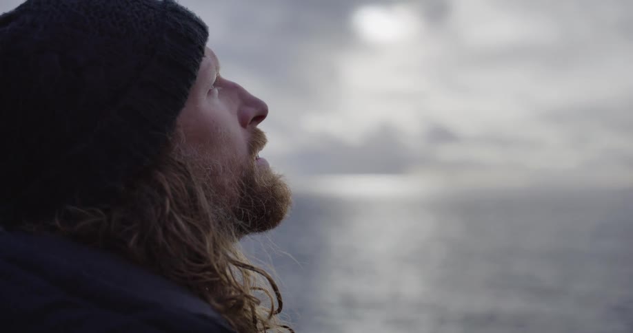 A slow-motion close-up of a contemplative man with a beard and long hair breathing in the crisp air as he looks out over a tranquil ocean