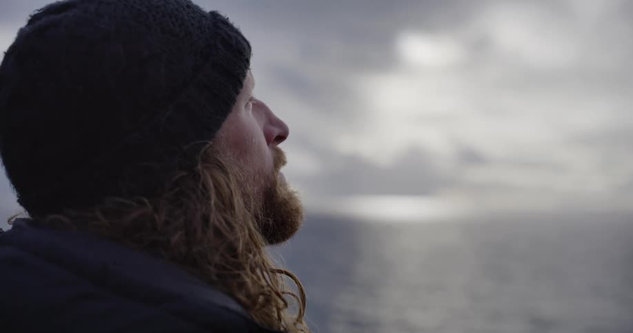 A contemplative man with a long beard and beanie gazes out at the shimmering ocean under a cloudy sky