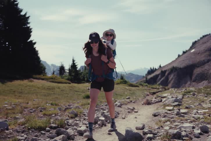A smiling mother carries her young child in a carrier backpack while hiking on a beautiful sunny day in the mountains