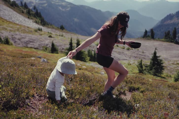 A mother holds her young child's hand as they forage for wild berries in a beautiful mountain meadow in slow motion