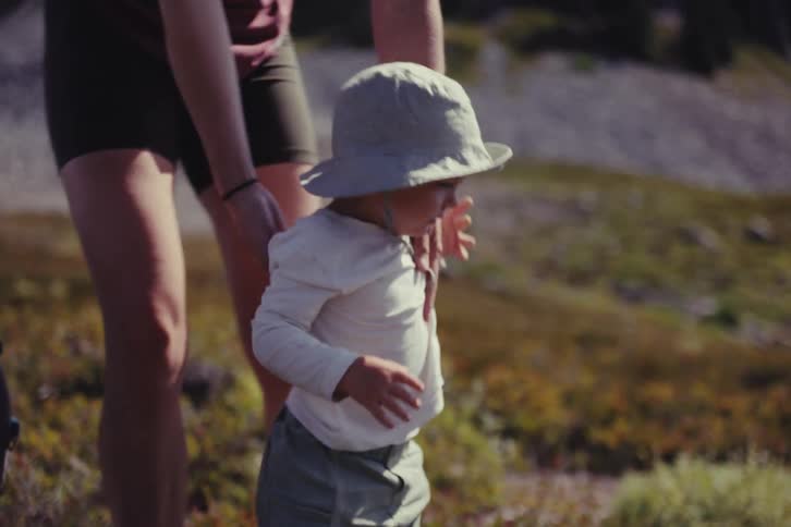 A mother lifts her crying toddler out of a hiking carrier pack and watches them walk along a mountain trail in slow motion