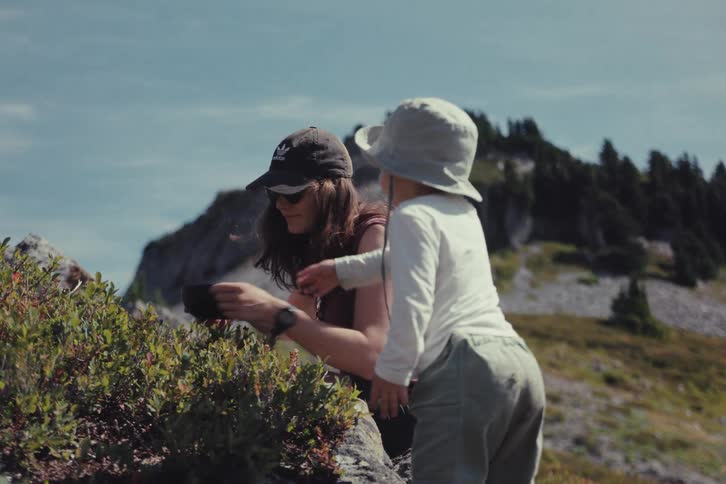 A mother and her toddler pick and eat wild berries together on a sunny mountain hike in slow motion
