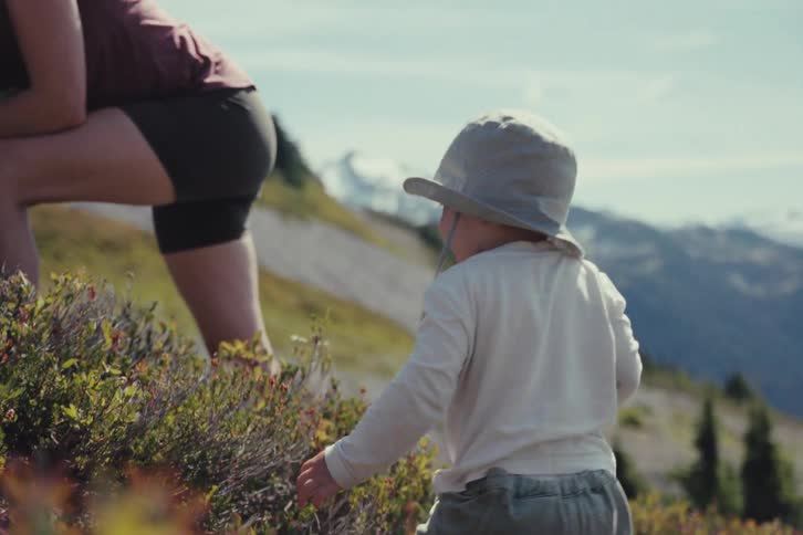 A happy toddler in a sun hat excitedly eats wild berries while hiking with his mother in the mountains in slow motion