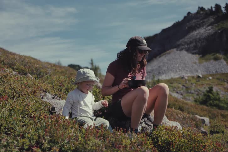 A mother and her young child sit on a sunny mountainside, taking a break to pick and eat fresh berries together