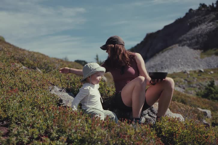 A mother and her toddler enjoy a sunny day in the mountains, taking a break from their hike to pick and eat fresh berries together