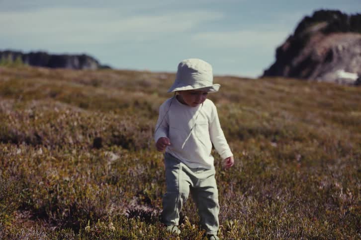 A cute toddler wearing a bucket hat happily walks through a vast mountain meadow in slow motion, exploring the beautiful landscape