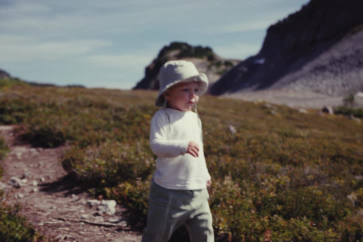 A cute toddler wearing a sun hat confidently explores a rugged mountain path on a beautiful sunny day