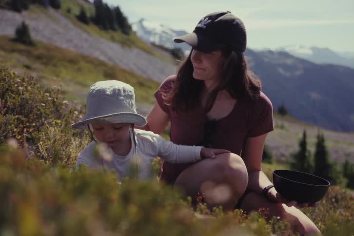 A young mother and her toddler enjoy a sunny day picking berries in an alpine meadow with snow-capped mountains in the distance
