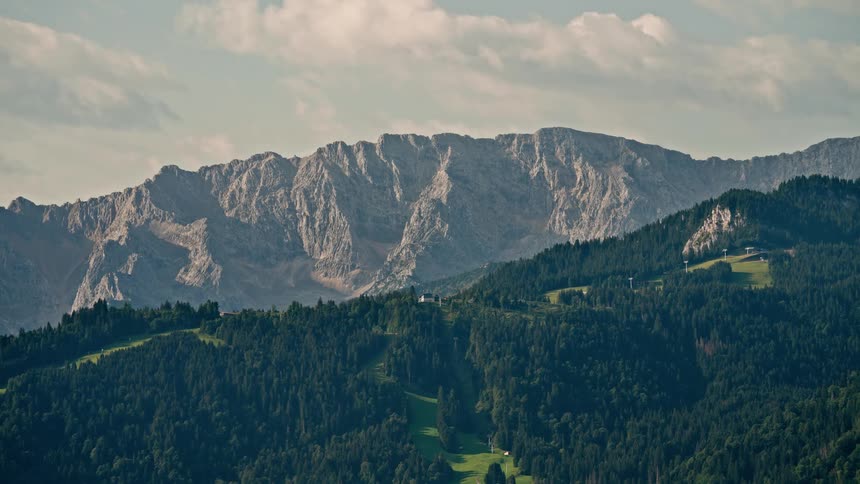 A majestic rocky mountain range towers over a lush green forested valley under a partly cloudy sky