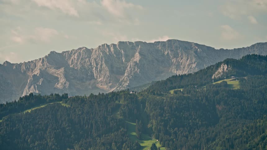 A serene time-lapse captures clouds moving over a majestic rocky mountain range and lush green forested hills