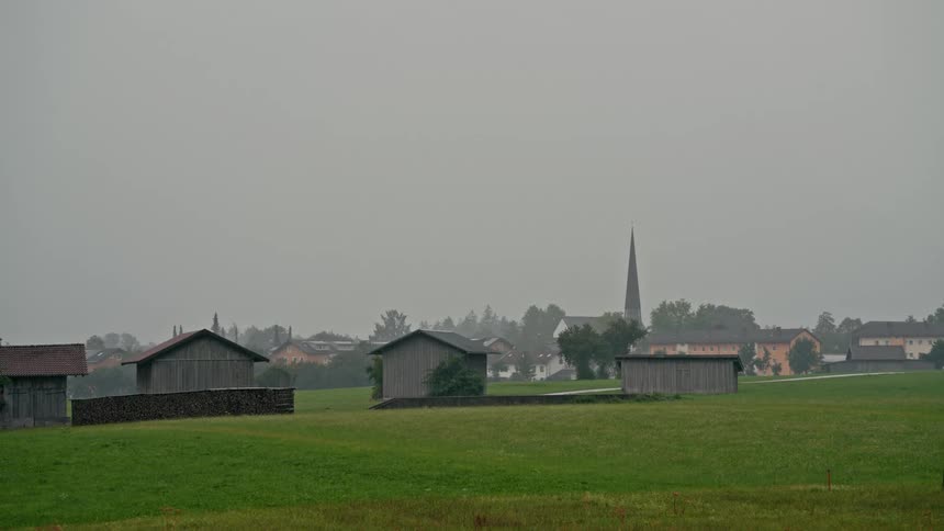 A quaint European village with a prominent church steeple sits in the distance behind a green pasture on a foggy day