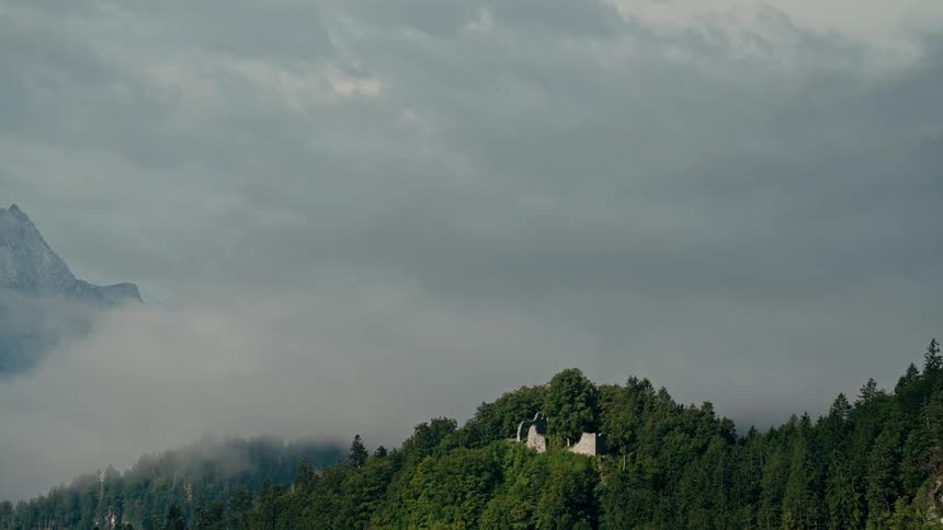 Timelapse of moody clouds and fog rolling over a forested mountain with ancient castle ruins