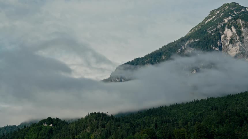 A majestic timelapse shows clouds and mist flowing over the green forested slopes of an alpine mountain