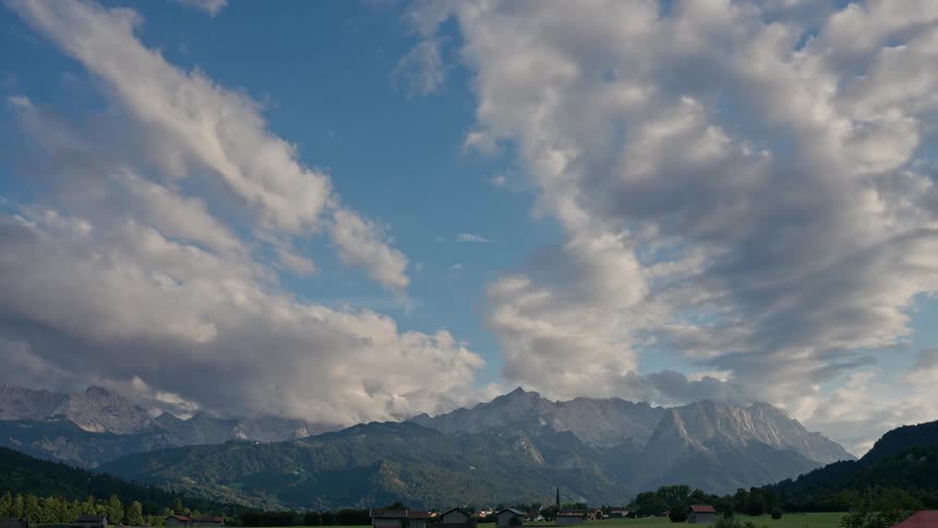 A serene timelapse shows fluffy clouds drifting across a vibrant blue sky above a majestic mountain range and a peaceful valley