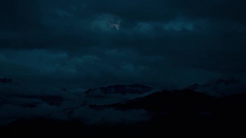 A faint moon emerges from behind dramatic dark clouds moving over a foggy mountain range at night