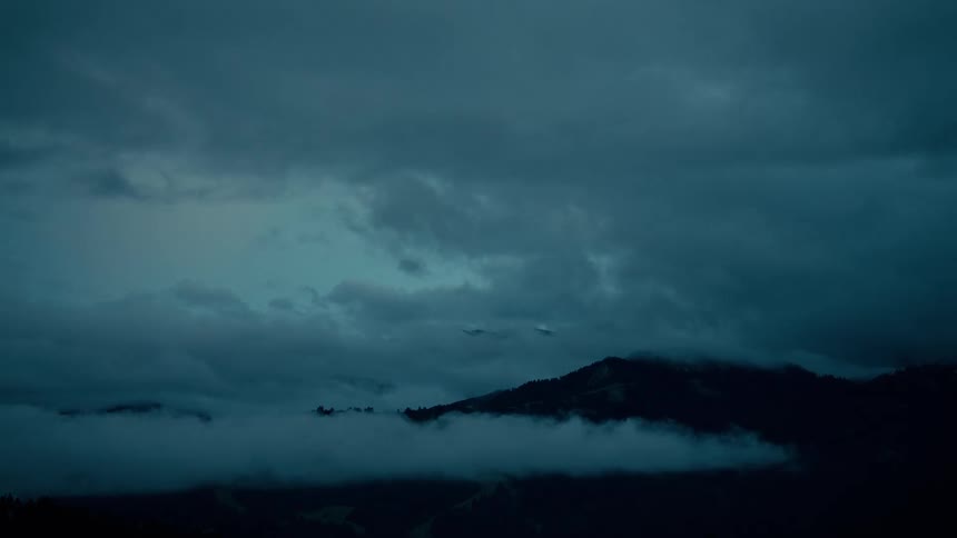 A moody timelapse shows the moon peaking through dense clouds moving over a mountain range from dusk to night