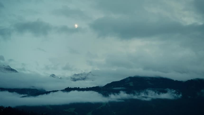 Mysterious clouds and fog drift over a dark mountain landscape as the moon slowly emerges in the gloomy sky