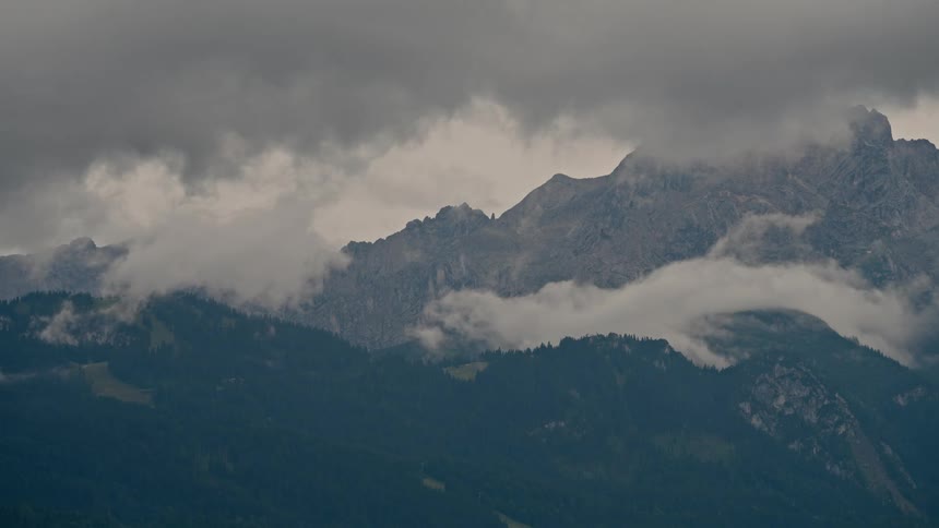 Gloomy, dark storm clouds and fog roll over a rocky mountain range covered in evergreen forests