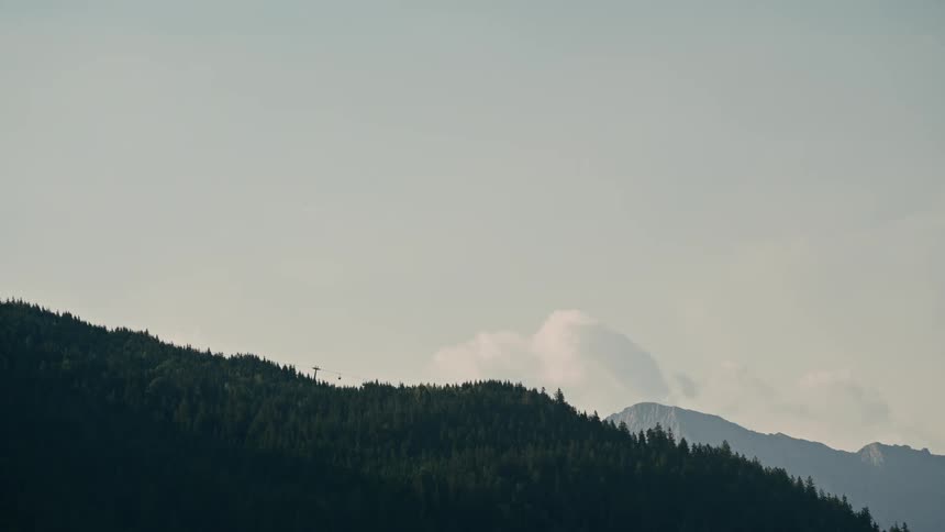 A lone gondola car slowly traverses the lush green forest of a mountain ridge under a hazy sky