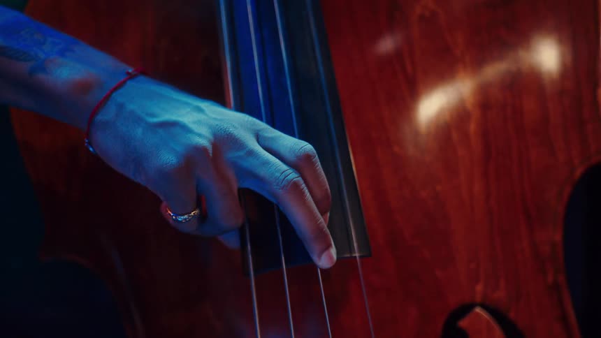 A close-up of a musician's tattooed hand playing the strings of a double bass under moody blue and red stage lighting