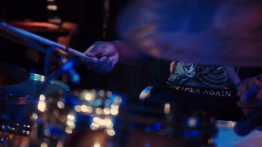 Slow motion close-up of a drummer's hands playing a steady beat on a drum kit under atmospheric blue stage lighting