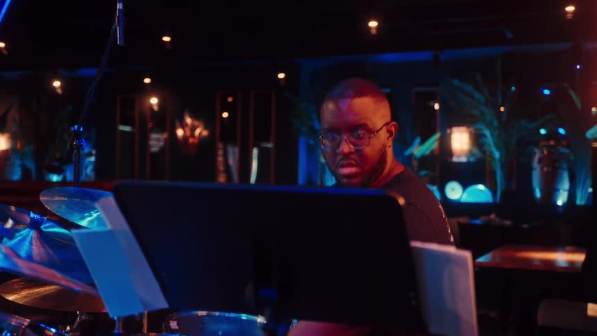 A talented male drummer with glasses plays a drum kit on a dimly lit stage during a live musical performance