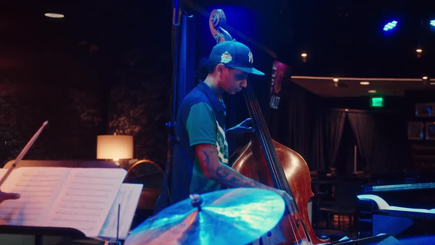 A tattooed musician wearing a baseball cap plays his upright bass on a dimly lit stage at a jazz club