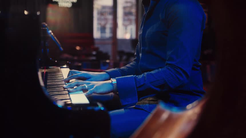 A smiling jazz pianist performs on a grand piano in a moody, blue-lit music club, with the camera moving from his hands to his happy face