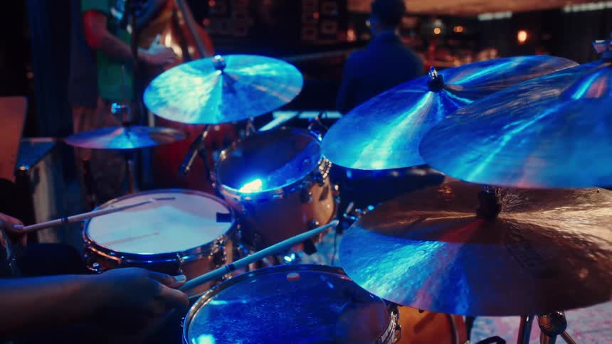 A musician plays the drums with a band during a live performance in a club bathed in cool blue light
