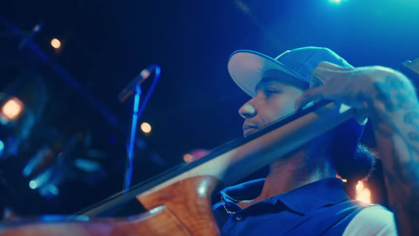 A stylish musician with tattoos passionately plays the upright bass during a live performance under vibrant blue stage lights