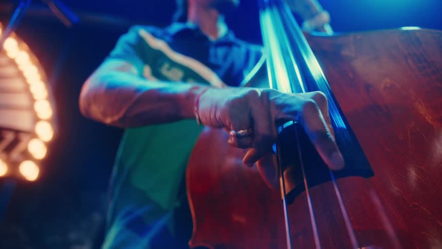 A musician's hands slide and pluck the strings of a wooden double bass during a live performance in a moody, colorfully lit club