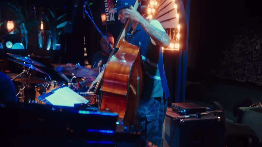 The reflection of a bass player on a grand piano slowly reveals a live jazz band performance in a dimly lit club