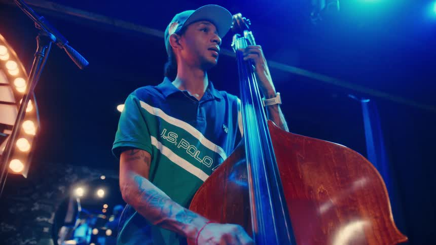 A jazz duo with a pianist and bassist performs on a dimly lit stage, with the camera focusing on the bassist playing his upright bass