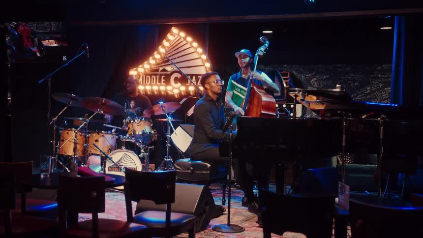 A jazz trio with a pianist, bassist, and drummer performs on a warmly lit stage in an empty club