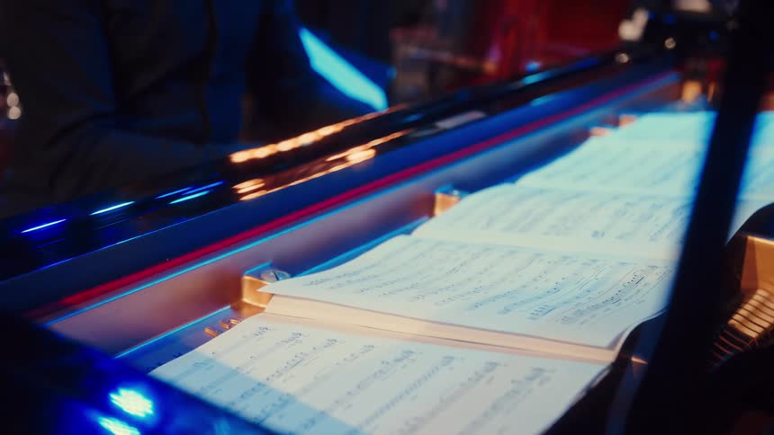 A musician's hands turn the page of sheet music while playing a grand piano under moody blue stage lighting