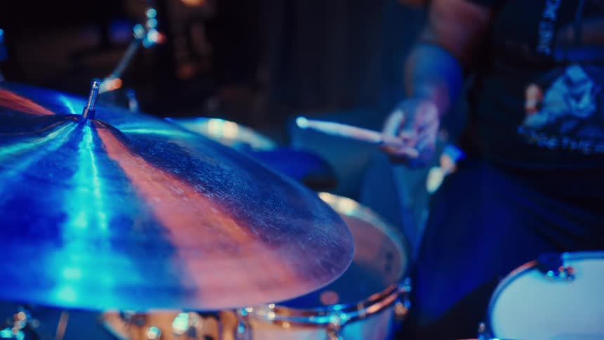A drummer plays his set in slow motion on a dimly lit stage, with a focus pull from the cymbal to his face