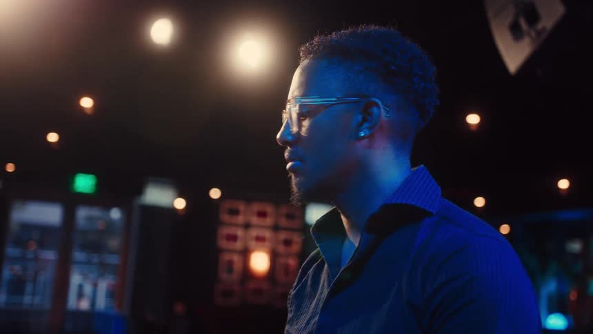 A focused man with glasses begins to play a grand piano on a dimly lit stage, captured in a slow-motion profile shot with cool blue lighting
