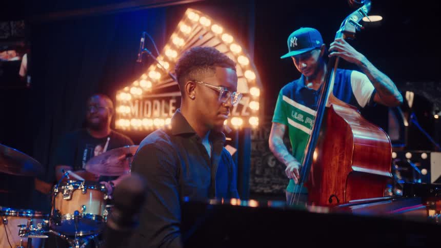 A talented jazz trio performs a soulful set on a dimly lit stage, with the pianist in the foreground and the drummer and bassist in the background