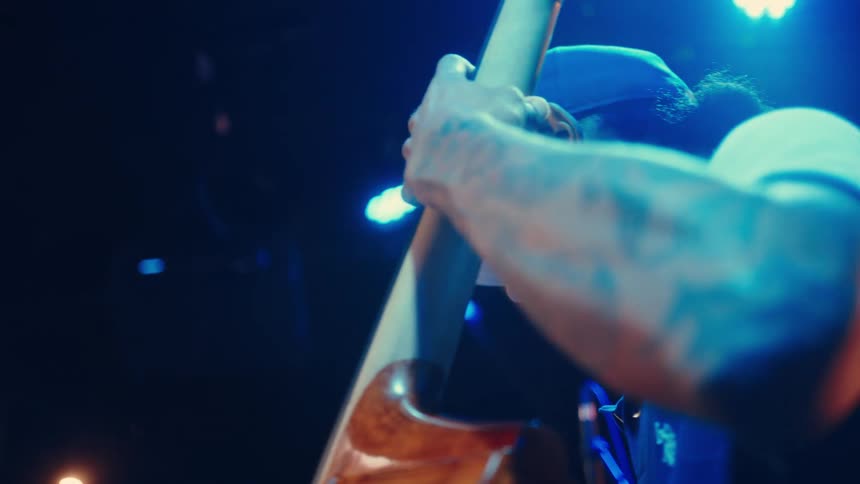 A tattooed musician passionately plays the upright bass in slow motion under dramatic blue stage lighting