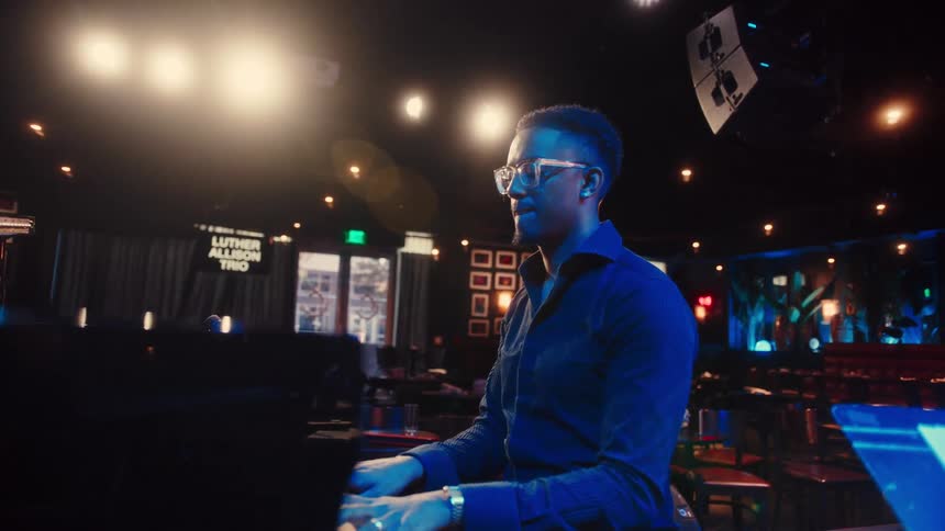 A stylish musician plays a grand piano on stage during a rehearsal in a moody, empty jazz club