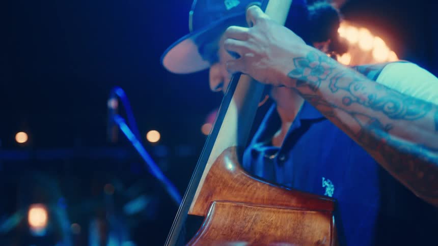 From the sheet music to the musician, a tattooed man in a cap plays the upright bass under cool blue stage lights