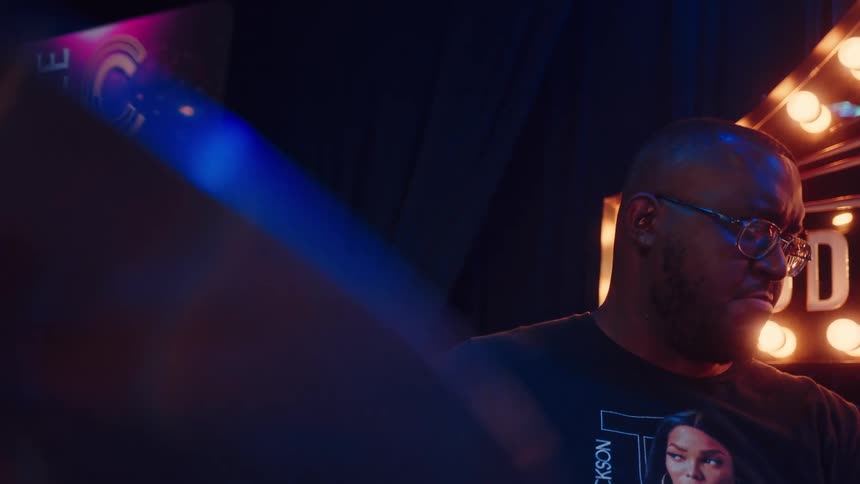 An intimate, slow-motion portrait of a drummer concentrating on stage, bathed in blue light with a lit sign behind him