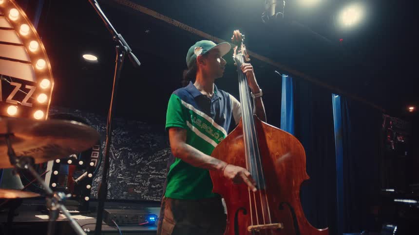 A cool bassist with long hair and tattoos plays the upright bass on stage at a dimly lit jazz club