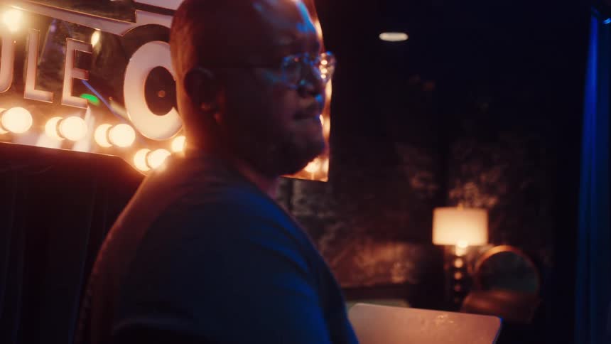 A jazz drummer with glasses passionately plays the drums on a dimly lit stage during a live performance, starting with an artistic rack focus