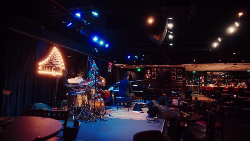 A jazz trio consisting of a pianist, drummer, and bassist performs together on a moody, blue-lit stage in a classic nightclub