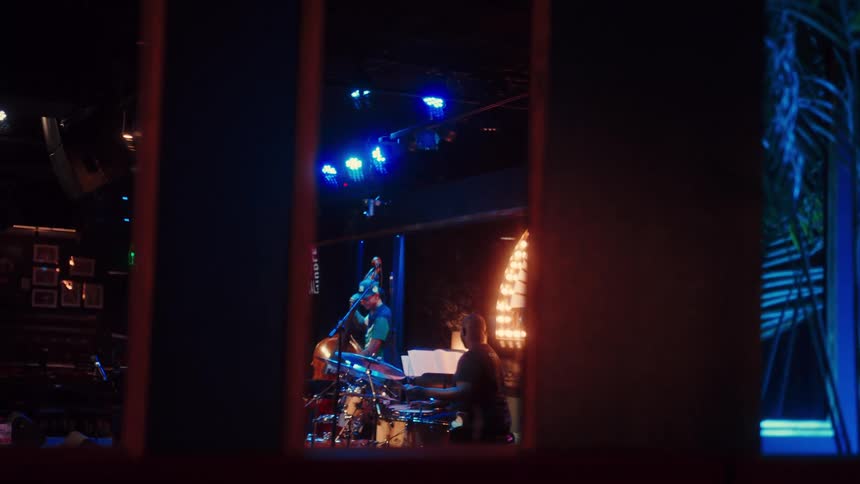 A jazz trio featuring a pianist, double bassist, and drummer performs on a dimly lit stage with blue lights, viewed from behind a window frame