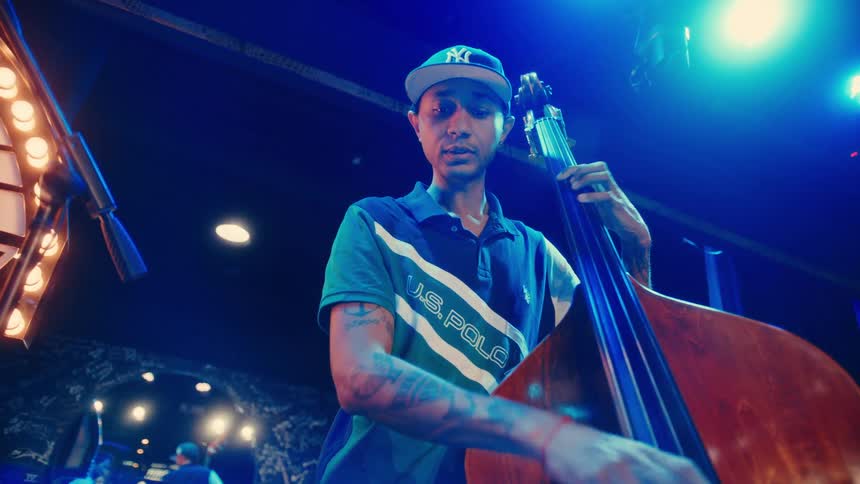 A tattooed musician in a baseball cap plays the upright bass on a blue-lit stage during a live performance